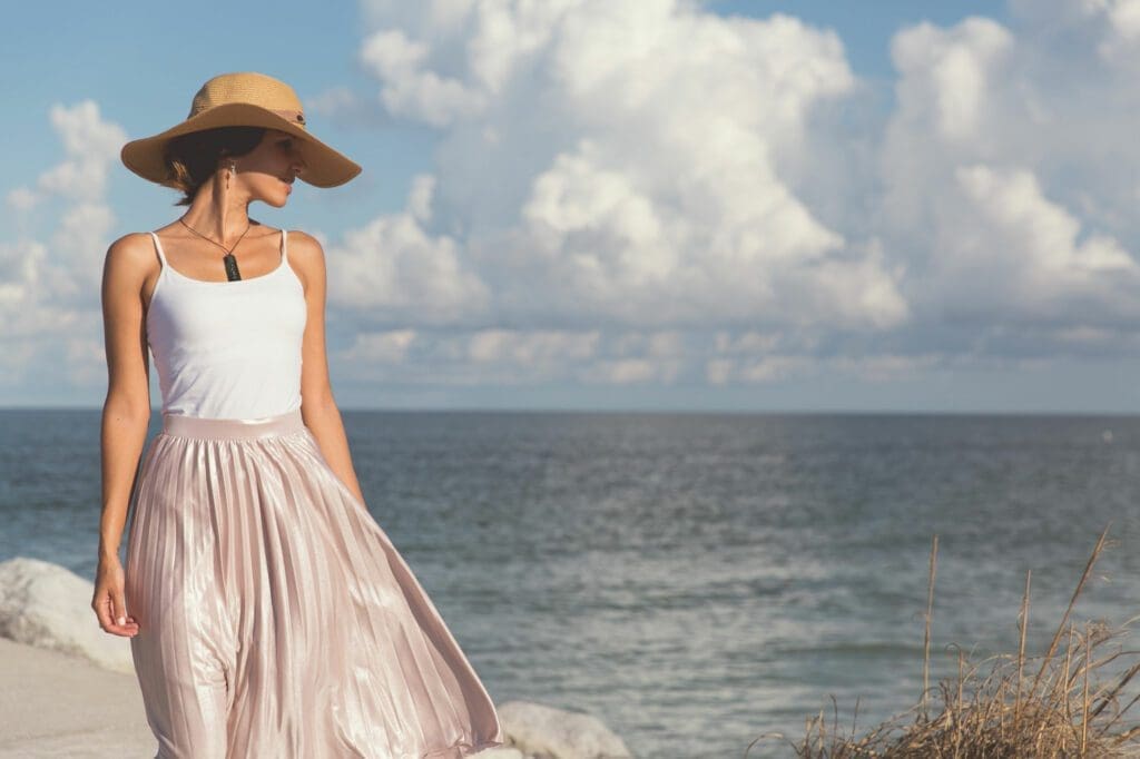 A woman in a hat and dress standing on the beach.