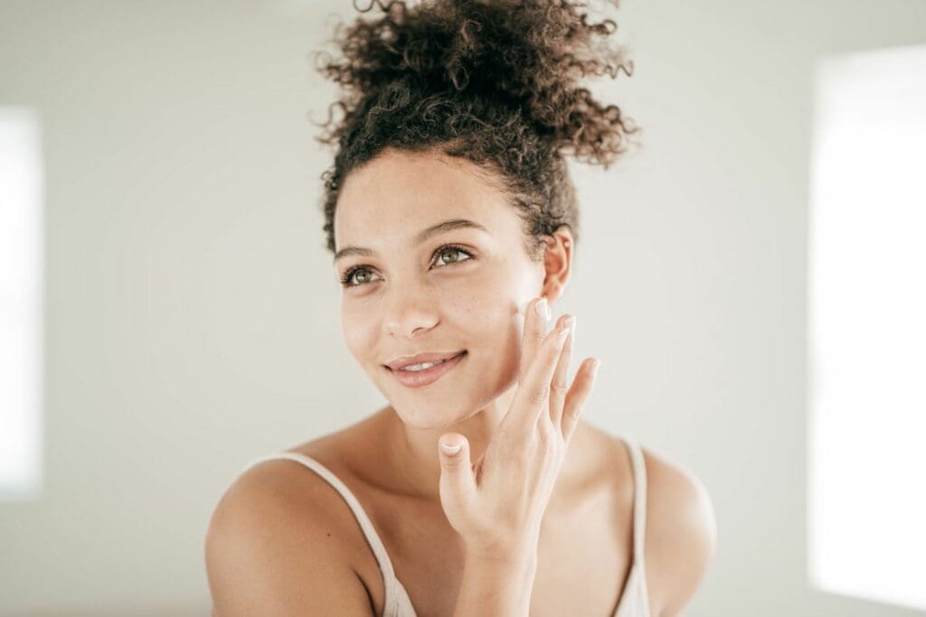 A woman with curly hair is smiling and touching her face.